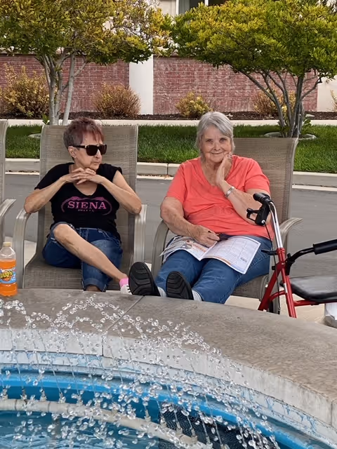 Two elderly women sitting on lounge chairs near a water fountain outdoors. One woman is wearing sunglasses and a black Siena t-shirt, while the other is in a coral shirt with a walker beside her, holding a newspaper. Trees and a brick wall are visible in the background.