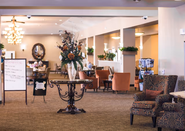 Spacious senior living common area with upholstered chairs, a central table holding a floral arrangement, and dining tables in the background.
