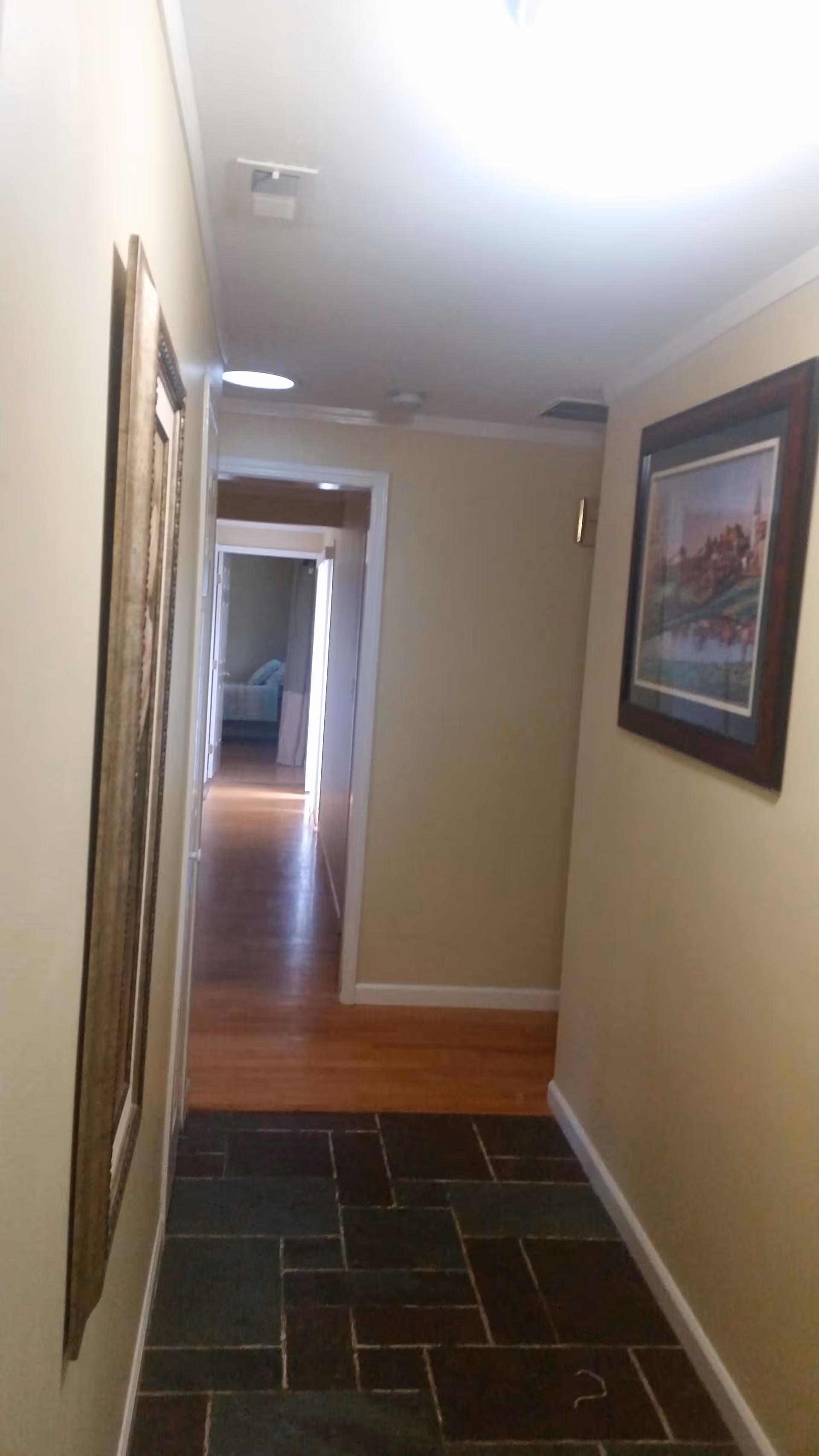 A narrow indoor hallway with beige walls and a dark tiled floor leading to a room with wooden flooring and a bed visible in the distance. The hallway has framed artwork on the walls and ceiling lights.