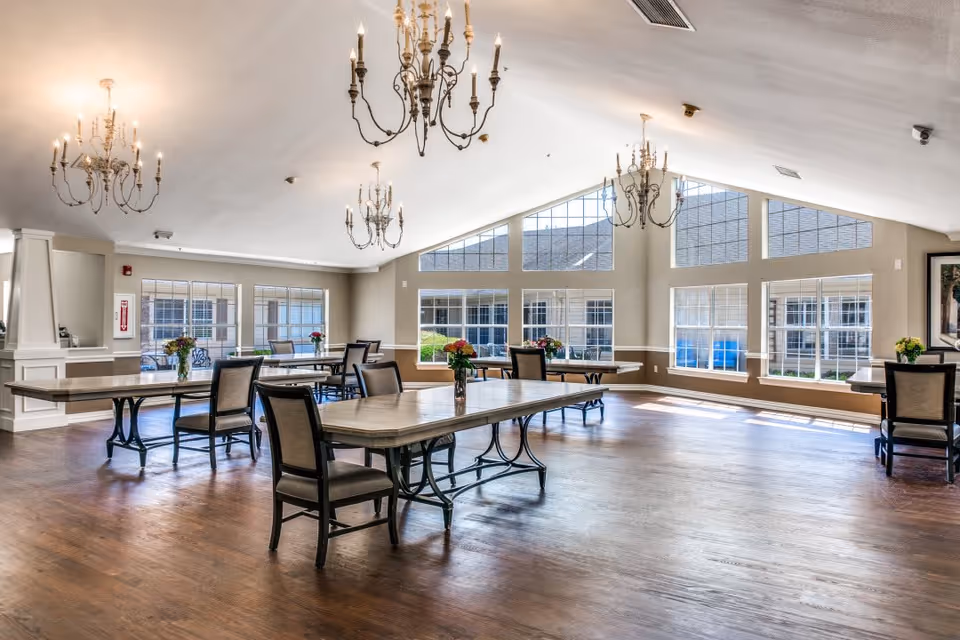 Spacious dining room with multiple tables and chairs, chandeliers, and large windows letting in natural light.