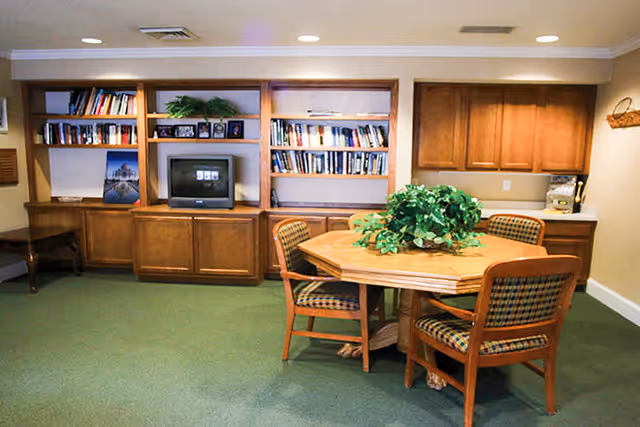 A cozy common area with a wooden octagonal table surrounded by four cushioned chairs with plaid upholstery. Behind the table is a built-in wooden shelving unit filled with books, framed photos, and a small television. To the right, there is a countertop with wooden cabinets above and below, and a coffee maker on the counter. The room has green carpet and beige walls with white crown molding.