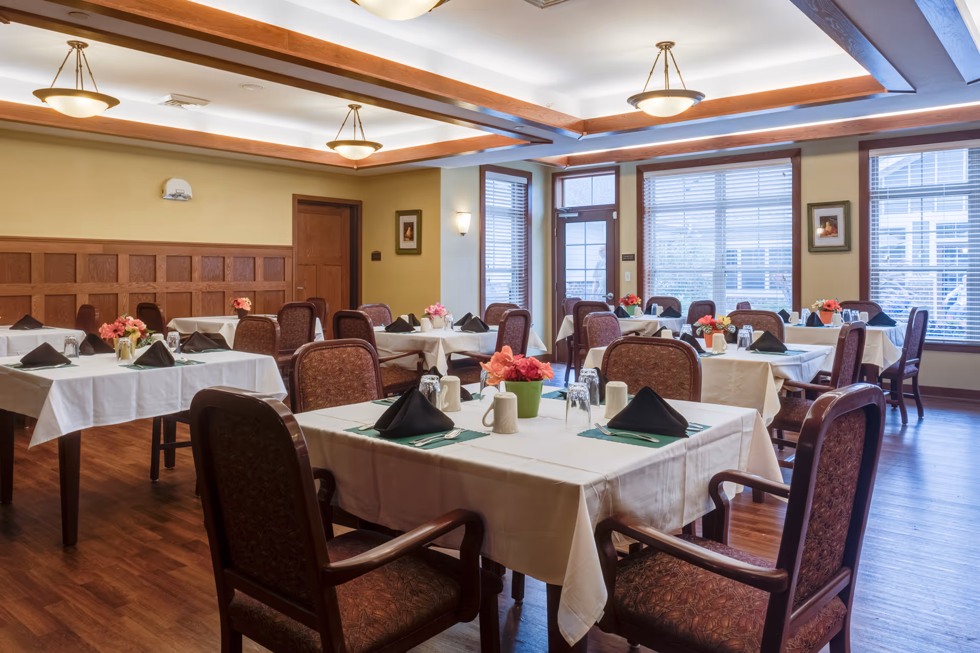 A dining room with multiple tables covered in white tablecloths, each set with black folded napkins, silverware, mugs, and glasses. The tables have small flower arrangements in the center. The room has wooden floors, beige walls with wood paneling, large windows with blinds, and ceiling lights with wooden beams.