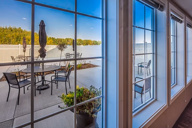 View through large windows showing an outdoor patio area with round tables, chairs, closed umbrellas, and some plants, surrounded by a white fence and trees in the background under a clear blue sky.