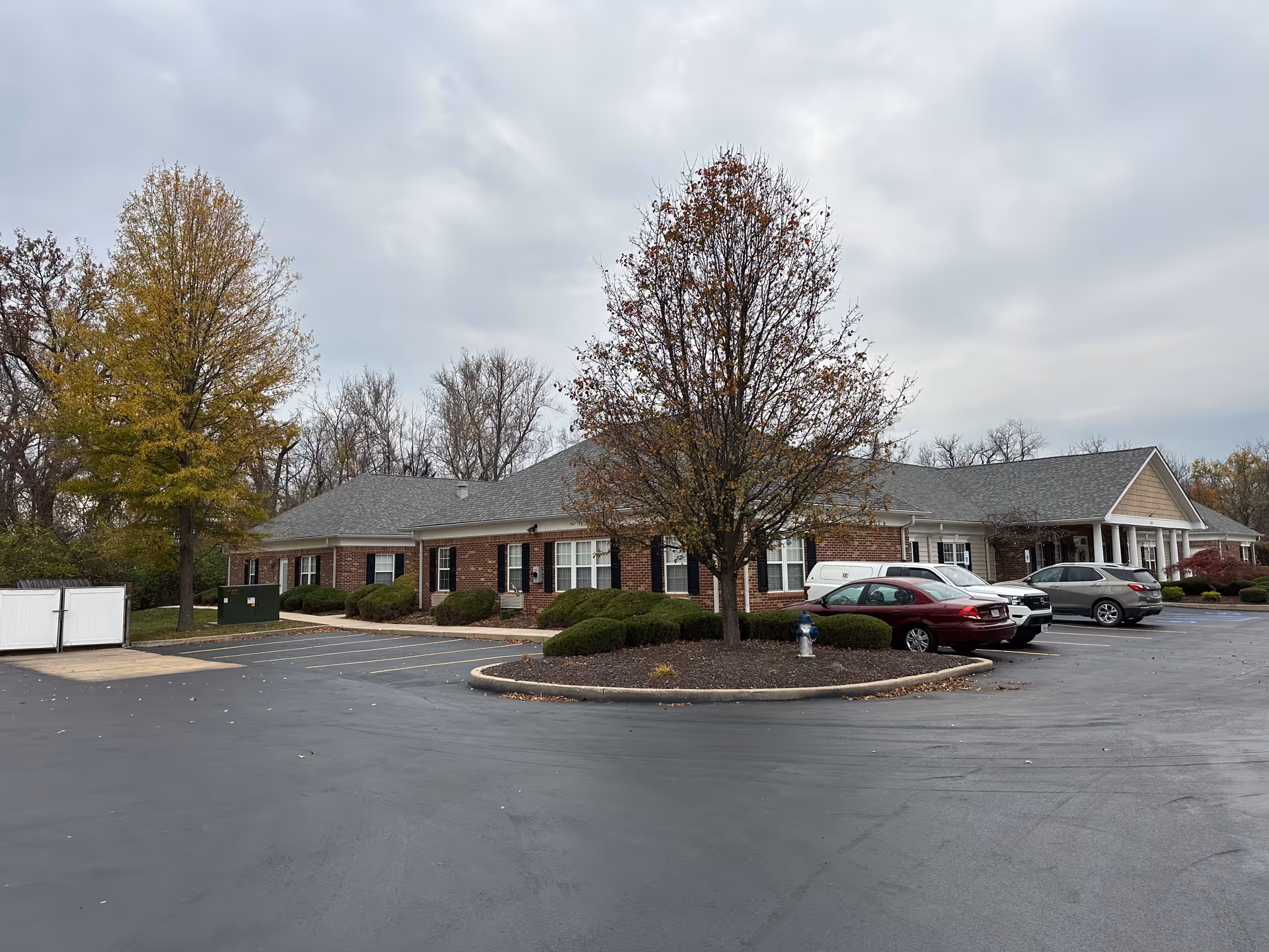 Exterior view of a single-story brick building with a gray roof, surrounded by a parking lot with several parked cars and landscaped areas with trees and bushes under a cloudy sky.