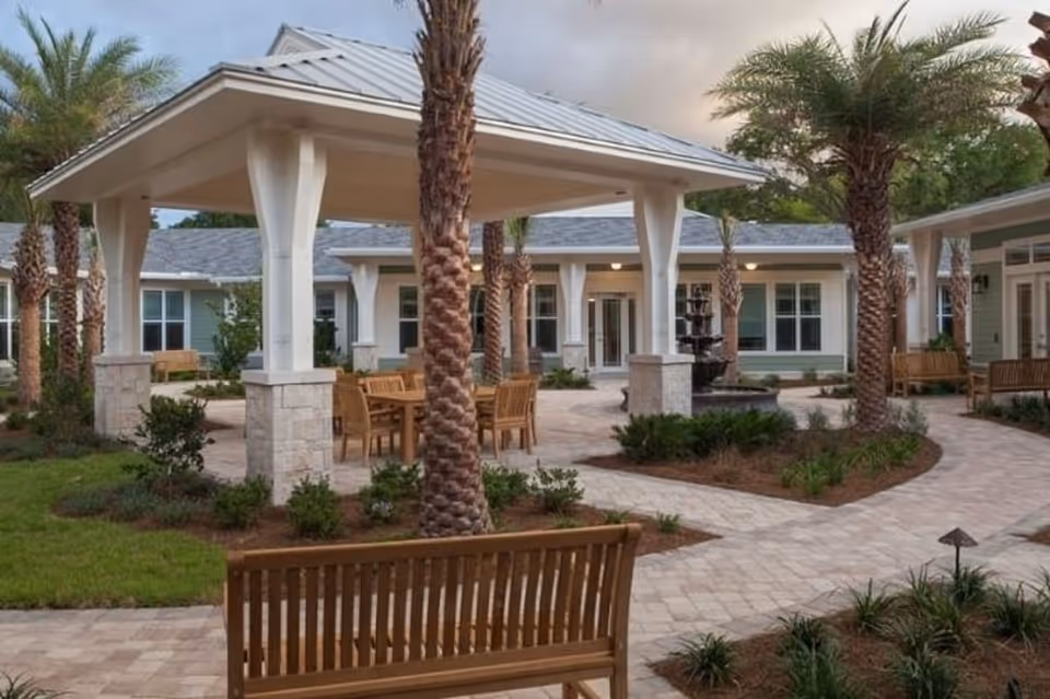 Outdoor courtyard area at Ponte Vedra Gardens featuring a covered seating area with wooden tables and chairs, surrounded by palm trees, landscaped plants, paved walkways, and benches.