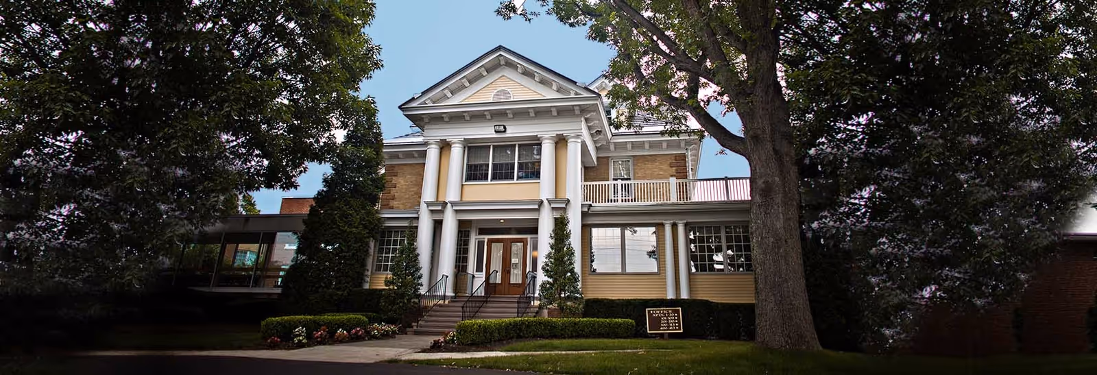 Front exterior of a two-story, columned senior living building with a central entrance, steps, landscaping, and large trees.