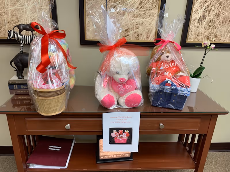 A wooden table with three gift baskets wrapped in clear cellophane and tied with red and pink ribbons. Each basket contains a stuffed animal and other items. Behind the table are framed pictures of wheat fields. On the table's lower shelf are books and a binder. A small potted orchid is on the right side of the table.