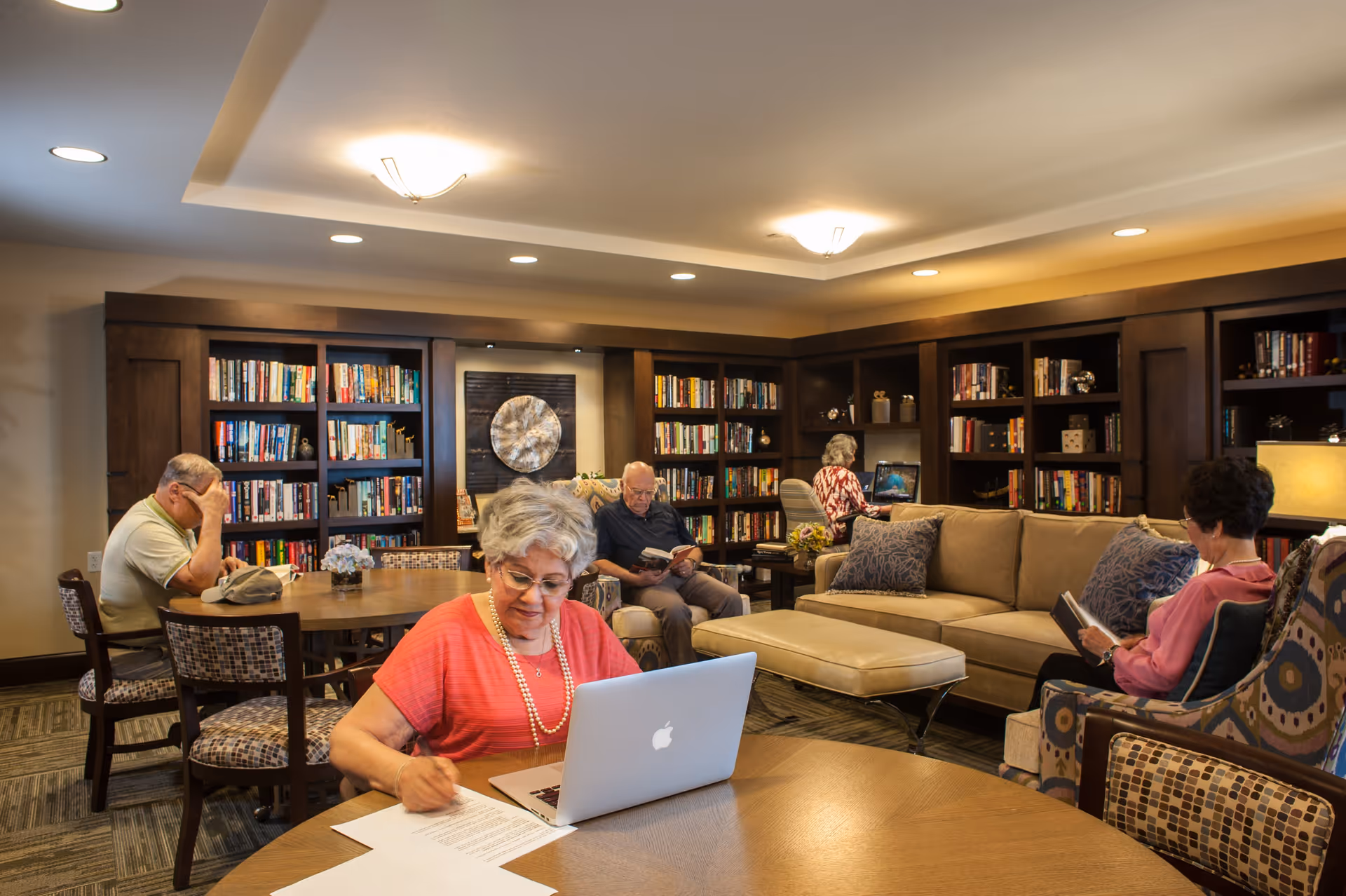A cozy library or common room in a senior living facility with several elderly people engaged in different activities. One woman in the foreground is working on a laptop and writing on papers at a round wooden table. In the background, a man is reading a book while sitting in a chair, another woman is using a computer at a desk, and another woman is reading a book while seated in a patterned armchair. The room features built-in dark wood bookshelves filled with books, a beige sofa with patterned pillows, and warm lighting from ceiling fixtures.
