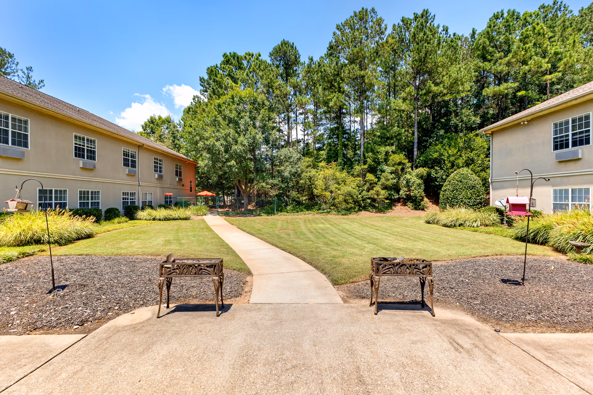 Outdoor courtyard area at The Reserve at Peachtree City Assisted Living and Memory Care featuring a concrete walkway leading through a grassy lawn with two beige two-story buildings on either side, surrounded by trees and landscaping under a blue sky with some clouds.