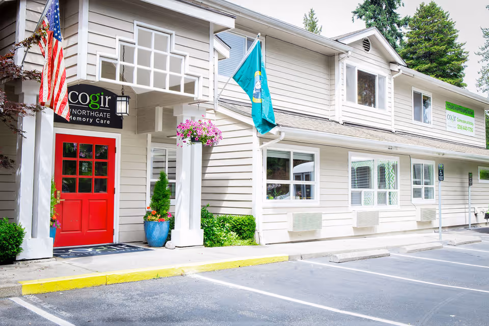 Exterior view of Cogir of Northgate Memory Care facility showing a beige building with a bright red door, American and Washington state flags, potted plants, and a parking area in front.