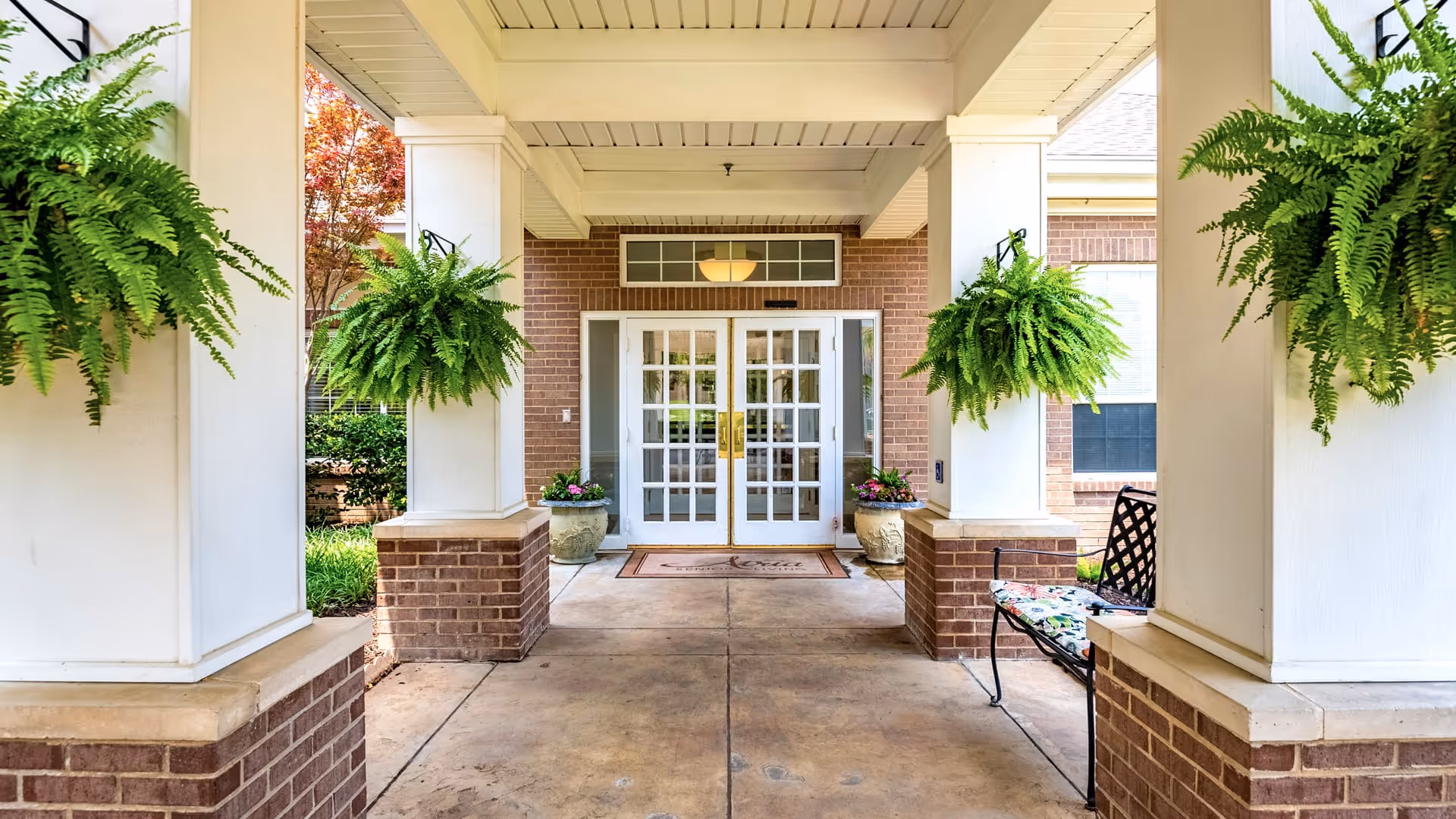 Covered entrance to a building with white double doors and glass panes, flanked by brick pillars topped with white columns. Four hanging green ferns are attached to the columns, and there are two large flower pots with colorful flowers near the doors. A bench with a floral cushion is placed on the right side. The area is clean and well-maintained with a concrete floor.