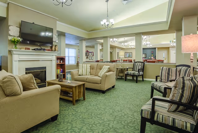 Communal living room with sofas and armchairs centered around a coffee table, a fireplace with TV above, and a dining area visible in the background.