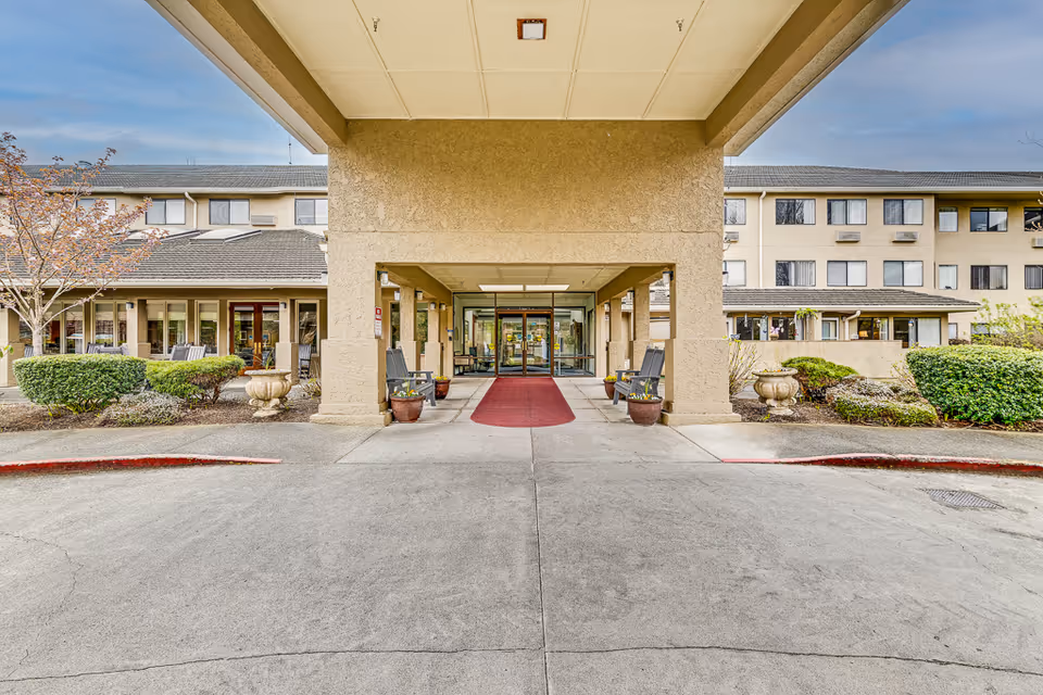 Covered main entrance and porte-cochère of a multi-story senior living building with a red carpet walkway, potted plants, and surrounding landscaping.
