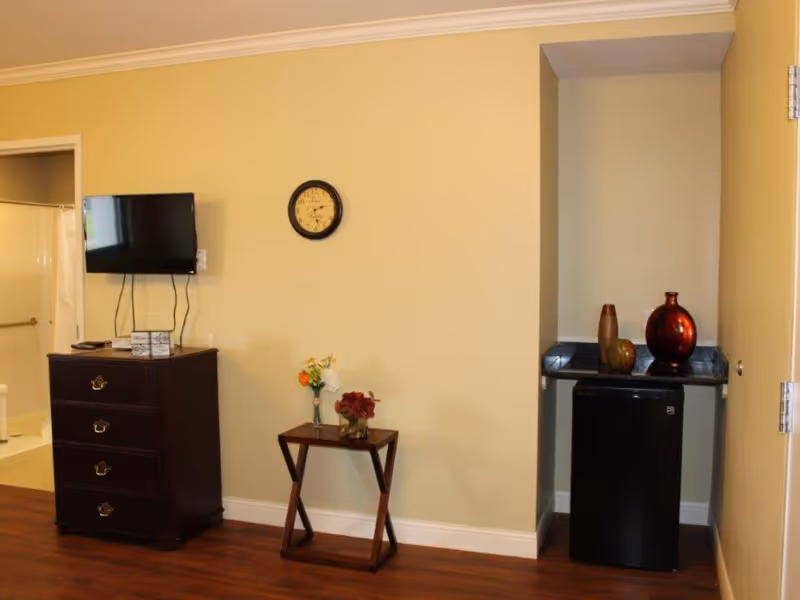 Interior room with a dresser and wall-mounted TV, a small table with flowers, and a mini-fridge in a recessed nook next to a bathroom doorway.
