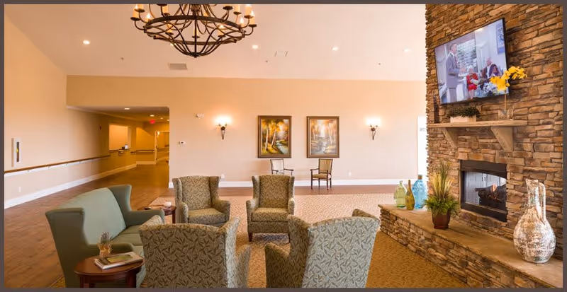 A cozy senior living lounge with upholstered chairs arranged around a stone fireplace and wall-mounted TV, a chandelier, and framed artwork on the far wall.