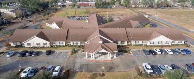 Aerial view of a single-story healthcare and rehabilitation center building with a brown roof and multiple wings extending from a central entrance. The building is surrounded by parking lots with several parked cars and some landscaped areas. The surrounding area includes open fields and residential neighborhoods.