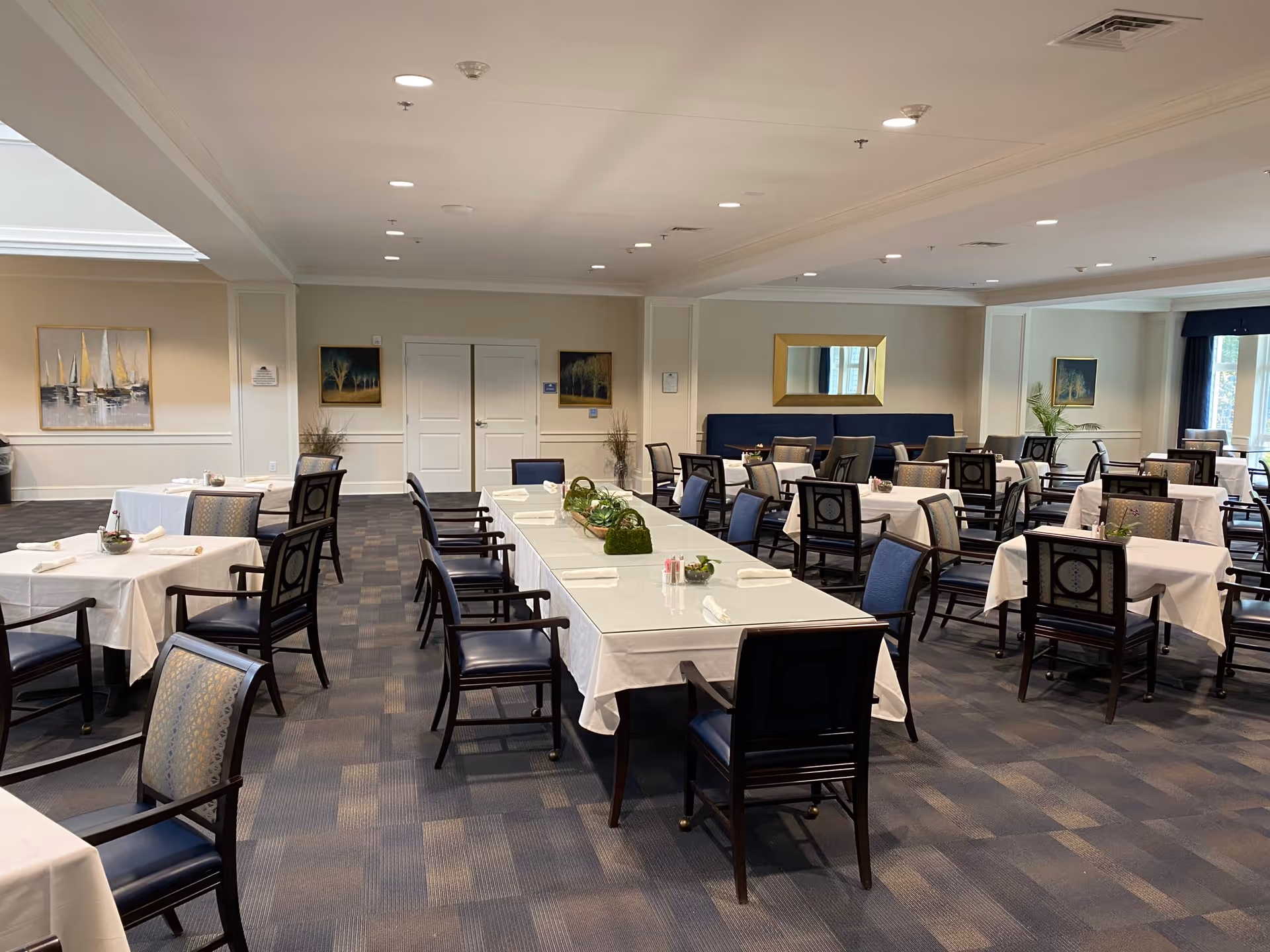 A spacious dining room with multiple tables covered in white tablecloths and surrounded by dark wooden chairs with blue cushions. The room has a carpeted floor, beige walls adorned with framed artwork, and a large mirror above a blue cushioned bench along one wall. The tables are set with napkins and small decorative plants, and the room is well-lit with ceiling lights and natural light from windows.