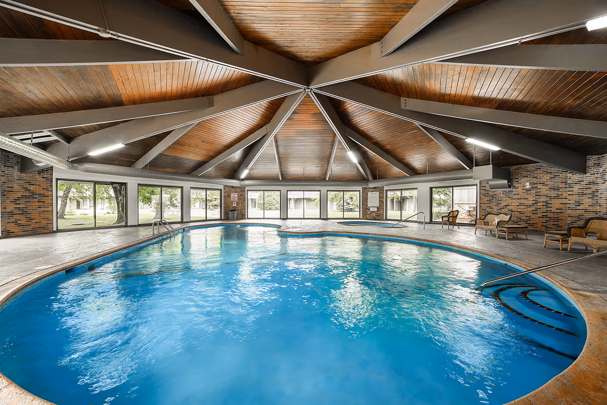 Indoor swimming pool with clear blue water under a wooden vaulted ceiling with exposed beams. Large windows surround the pool area, allowing natural light to enter and providing views of the outdoor greenery. Several wicker chairs and loungers are placed around the pool on the tiled floor.