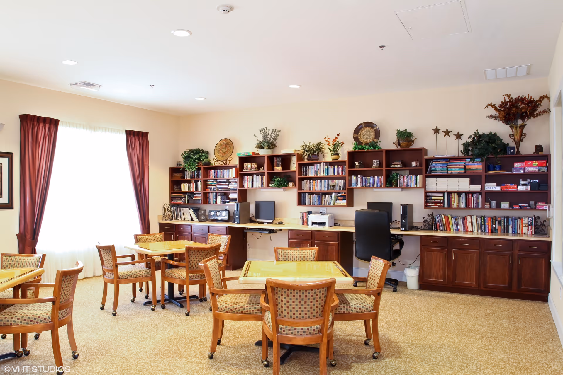 A bright room with several wooden tables and chairs arranged for group seating. The back wall features built-in wooden shelves filled with books, board games, and decorative plants. A computer and printer are also visible on the countertop beneath the shelves. Large windows with red curtains allow natural light to fill the space.