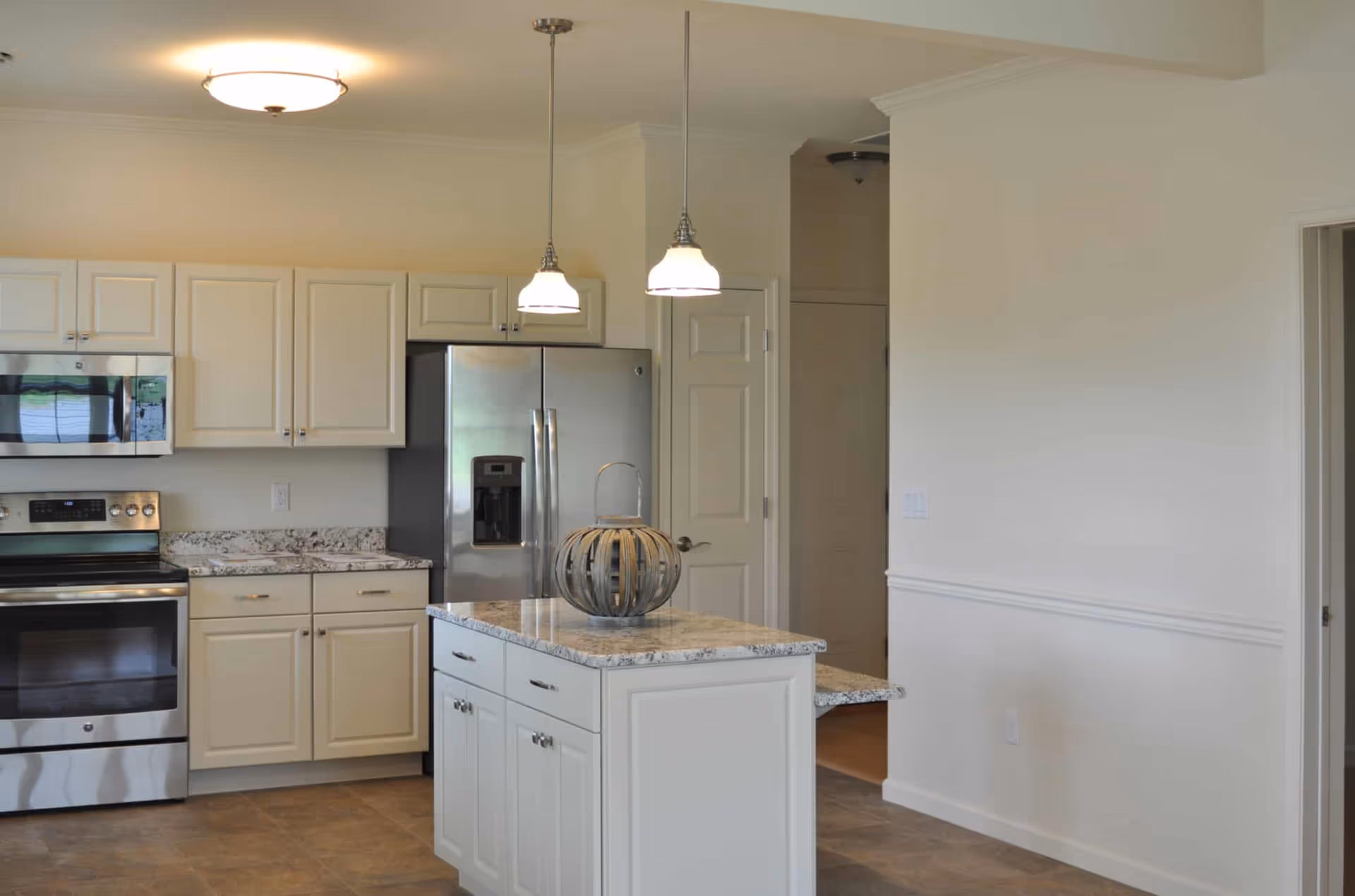 Modern kitchen with white cabinets, a stainless steel refrigerator, oven, and microwave. There is a kitchen island with a granite countertop and a decorative item on top. Two pendant lights hang above the island, and the floor is tiled.