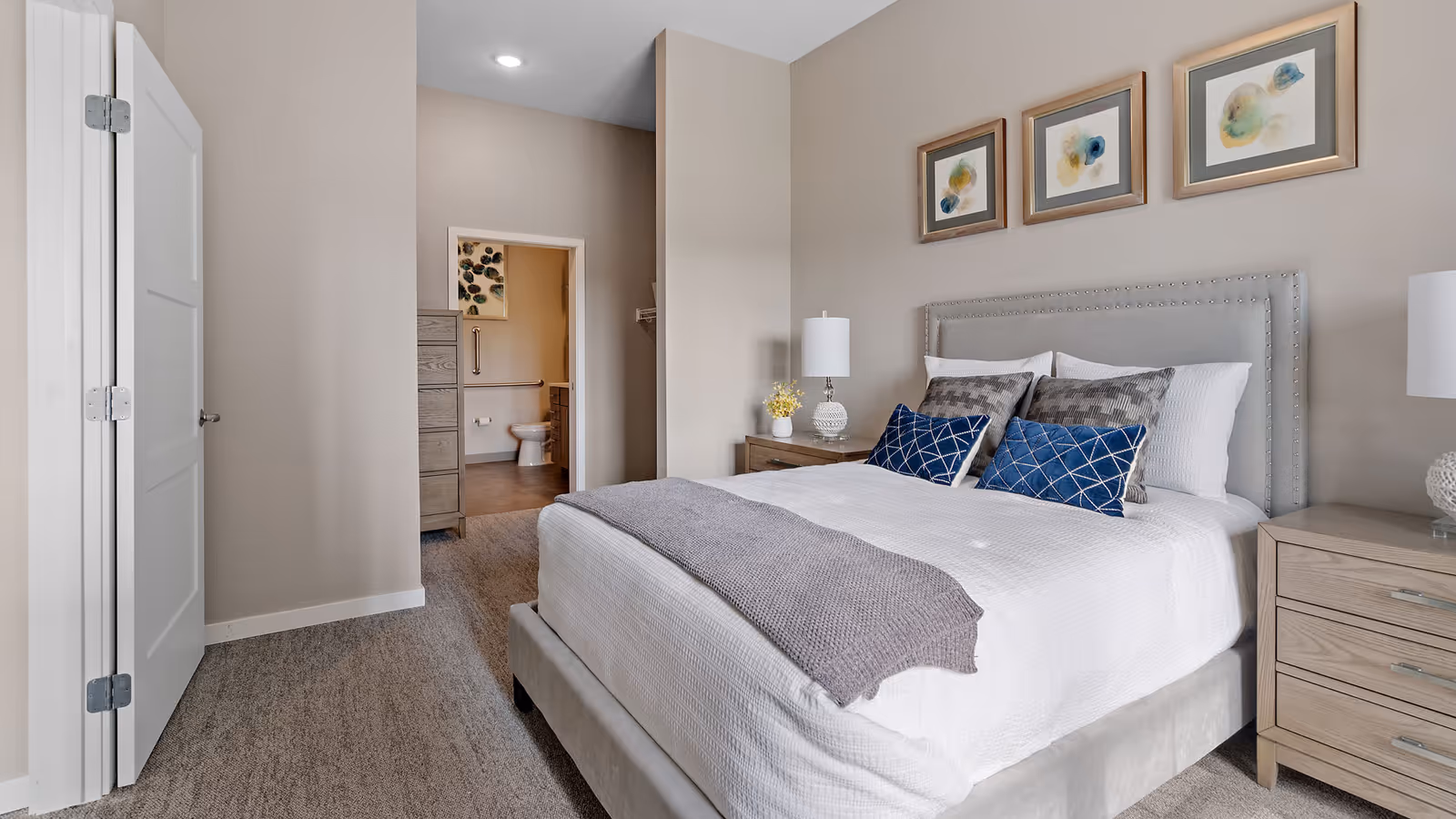 Well-lit bedroom with a bed, nightstands and lamps, decorative pillows, and an open door showing an en-suite bathroom.