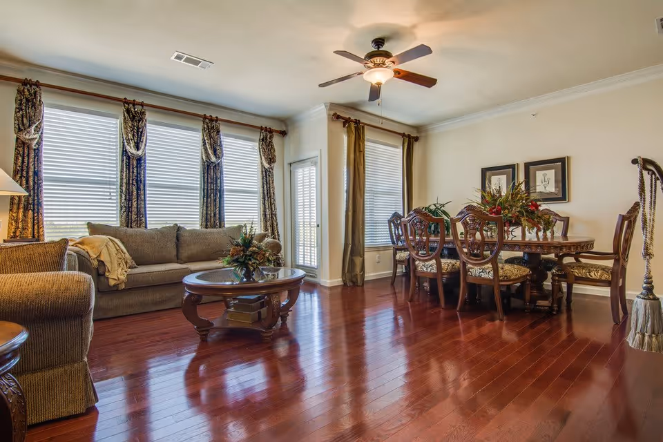 A bright living and dining area with polished wooden floors, a ceiling fan with light, a beige sofa with a throw blanket, a round wooden coffee table with a floral centerpiece, and a wooden dining table with six upholstered chairs. The room has large windows with blinds and decorative curtains, and framed artwork on the wall.