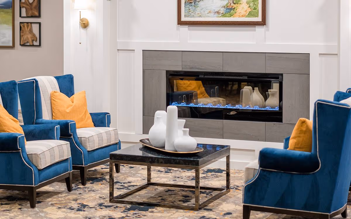 A cozy living room area with four blue upholstered armchairs featuring striped cushions and orange throw pillows arranged around a square black coffee table. On the table, there is a decorative tray holding white ceramic vases. Behind the seating area is a modern electric fireplace with a gray tiled surround, and above it hangs a framed landscape painting. The room has a patterned area rug and white walls with decorative molding.