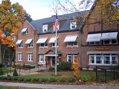 Exterior view of a brick building with multiple windows covered by white awnings. There is a flagpole with an American flag in front of the building, surrounded by a small garden with shrubs and trees showing autumn foliage. A sign on the building reads 'Southmayd Home.'