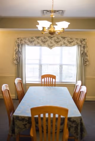 A dining room with a rectangular table covered by a blue patterned tablecloth surrounded by six wooden chairs. A large window with floral valance and curtains is in the background, and a brass chandelier with five lights hangs above the table.