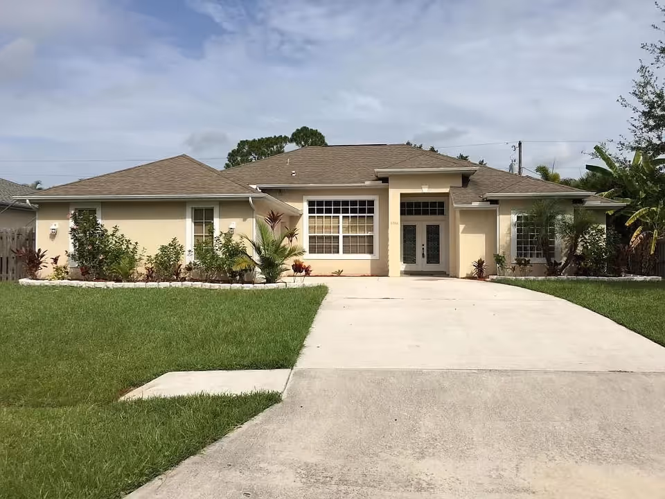Single-story beige assisted living building with a wide driveway, front lawn, large front windows, and landscaping under a cloudy sky.