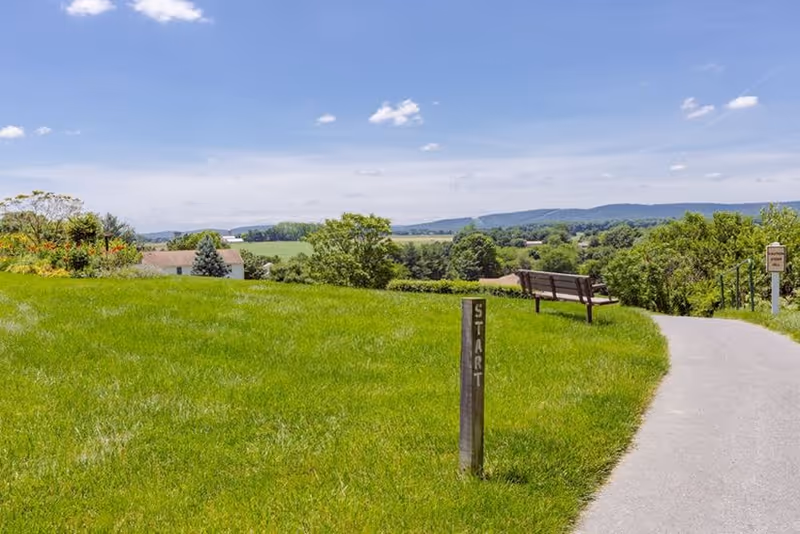 A paved walking path curves through a grassy area with a wooden post labeled 'START' and a bench nearby. Trees and bushes surround the area, with hills and a partly cloudy sky in the background.