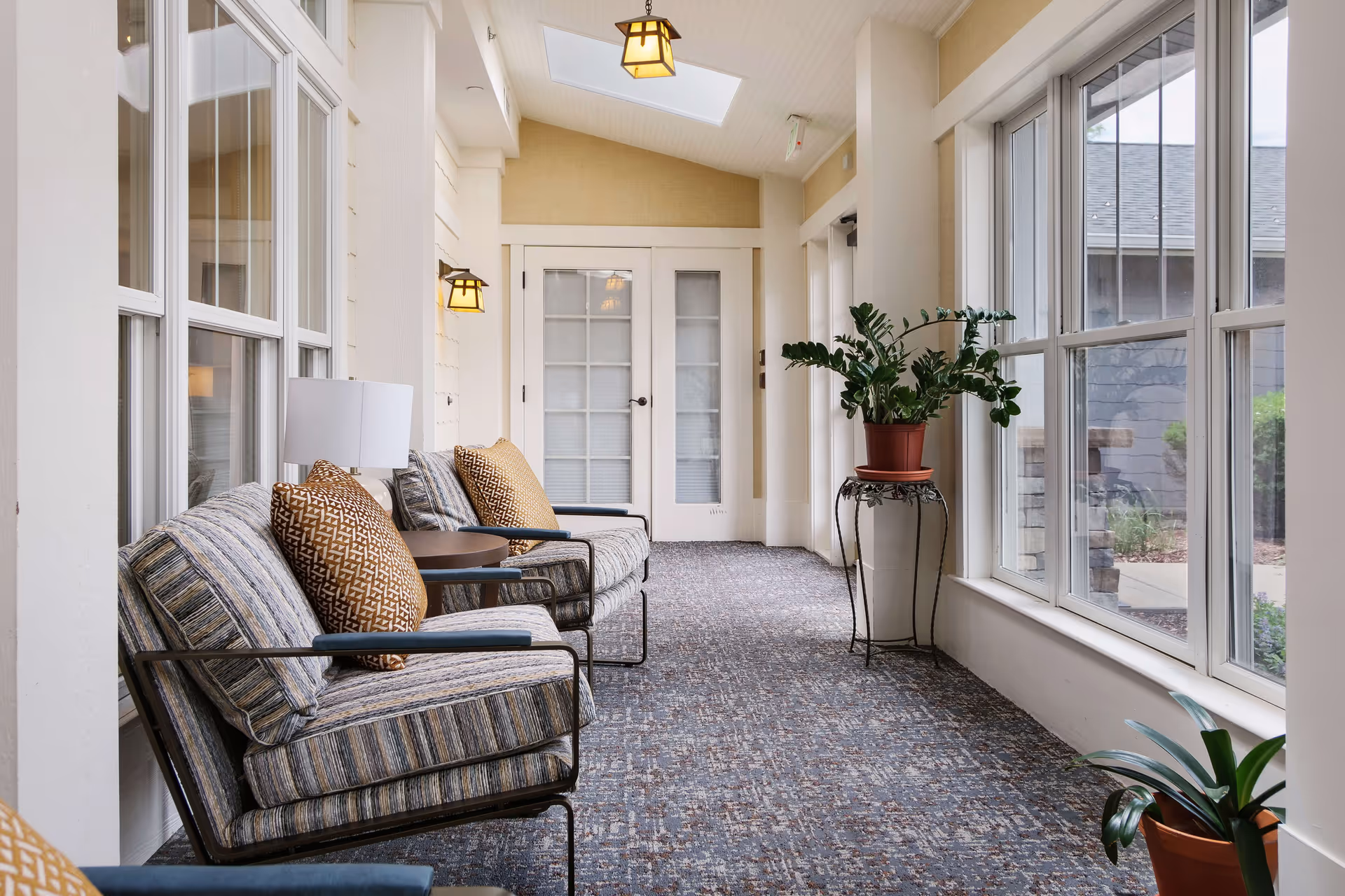 A bright indoor sitting area with large windows on the right side, featuring striped cushioned chairs with patterned pillows, small round tables, potted plants, and hanging lantern-style lights. The space has a carpeted floor and white walls with double glass doors at the end.