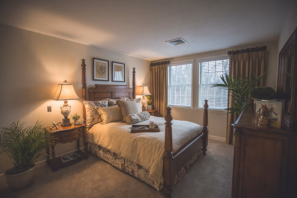 A cozy bedroom with a wooden four-poster bed adorned with multiple pillows and a tray with a cup on the bed. There are two matching wooden nightstands with lamps on either side of the bed, a potted plant on the left side, and large windows with beige curtains letting in natural light. A wooden dresser with decorative items and a large mirror is on the right side of the room.