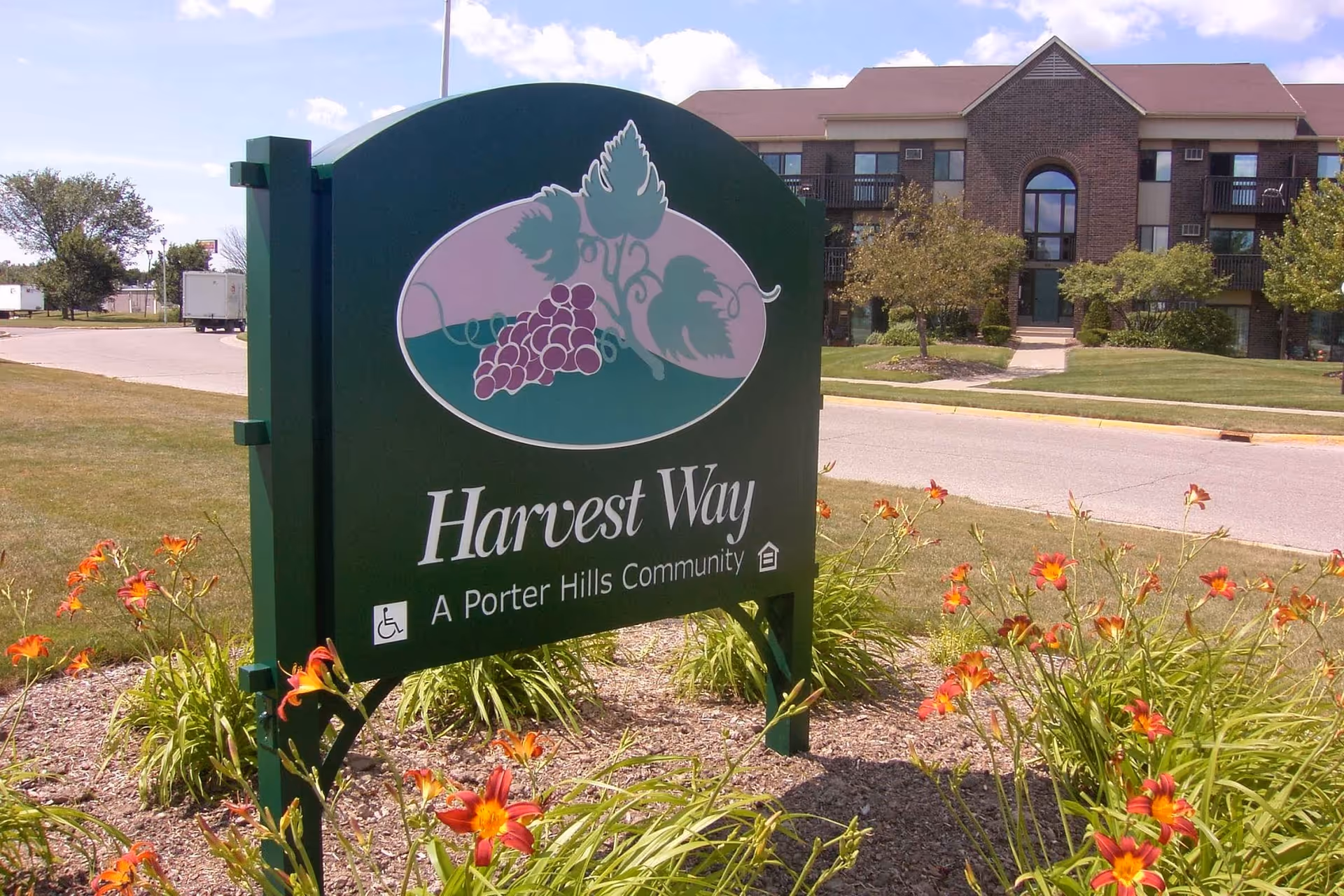 Outdoor view of a green sign for Harvest Way, a Porter Hills Community, surrounded by orange flowers and grass. In the background, there is a multi-story brick building with balconies and trees under a partly cloudy sky.
