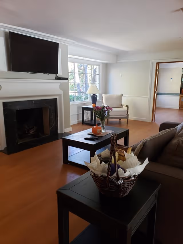 A cozy living room with a mounted flat-screen TV above a black marble fireplace. There is a large window letting in natural light, a white armchair next to a side table with a lamp, and a coffee table with a small pumpkin and a vase of flowers. In the foreground, there is a dark side table with a basket containing snacks and napkins. The room has wooden flooring and light-colored walls.