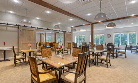 A spacious dining room with multiple wooden tables and chairs arranged neatly. The room features large windows allowing natural light to fill the space, pendant lights hanging from the ceiling, and sliding barn doors on one side. Small flower vases are placed on the tables, creating a welcoming atmosphere.