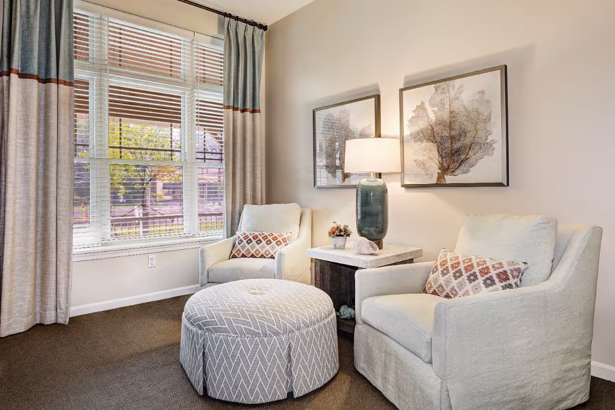 A cozy sitting area with two beige armchairs, each with a patterned cushion, positioned around a round patterned ottoman. Between the chairs is a small side table with a green ceramic lamp and a small plant. Two framed tree artworks hang on the wall behind the chairs. A large window with beige and blue curtains lets in natural light.