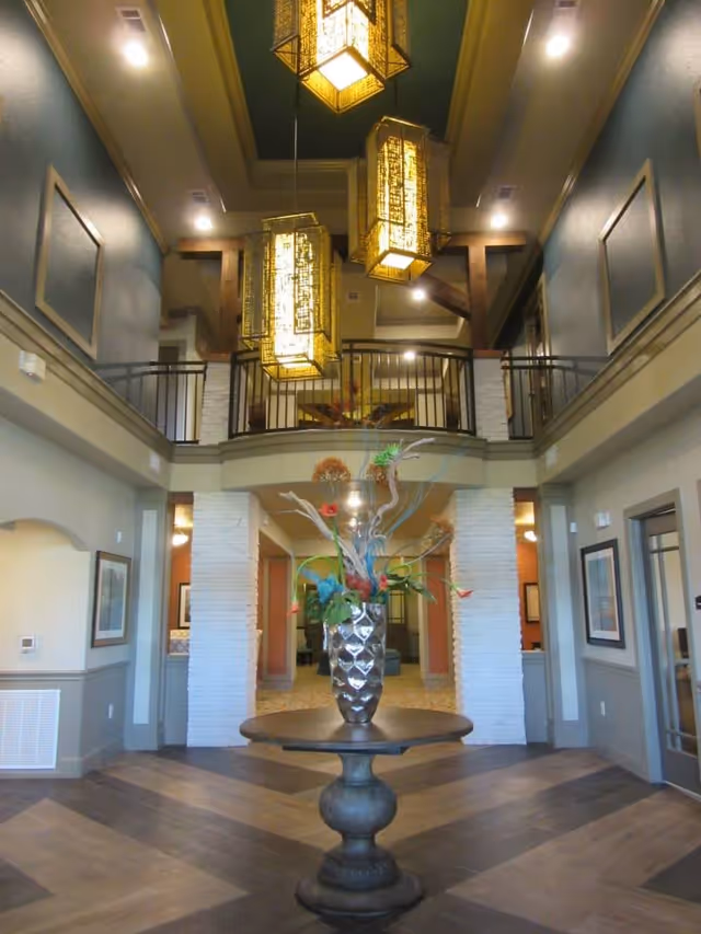Interior view of a senior living facility lobby with a round wooden table in the center holding a decorative vase with colorful flowers and branches. The space features high ceilings with three large hanging lantern-style lights, a second-floor balcony with black railings, and walls painted in muted tones with framed artwork.