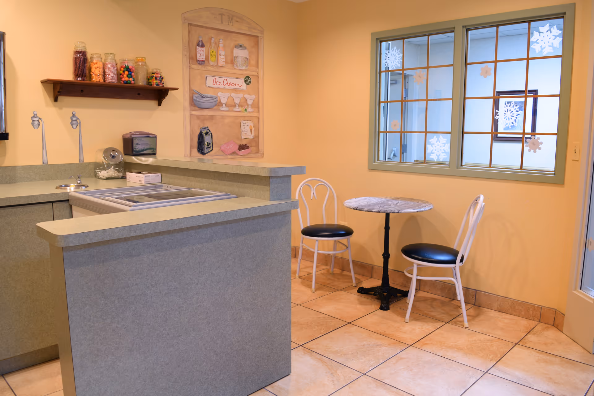 Small café-style dining area with a service counter, candy jars and ice-cream wall art, plus a round table and two chairs by a window.