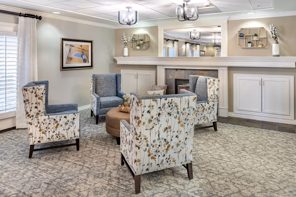 Cozy communal sitting room with patterned armchairs arranged around a round ottoman in front of a fireplace and mirrored mantel.
