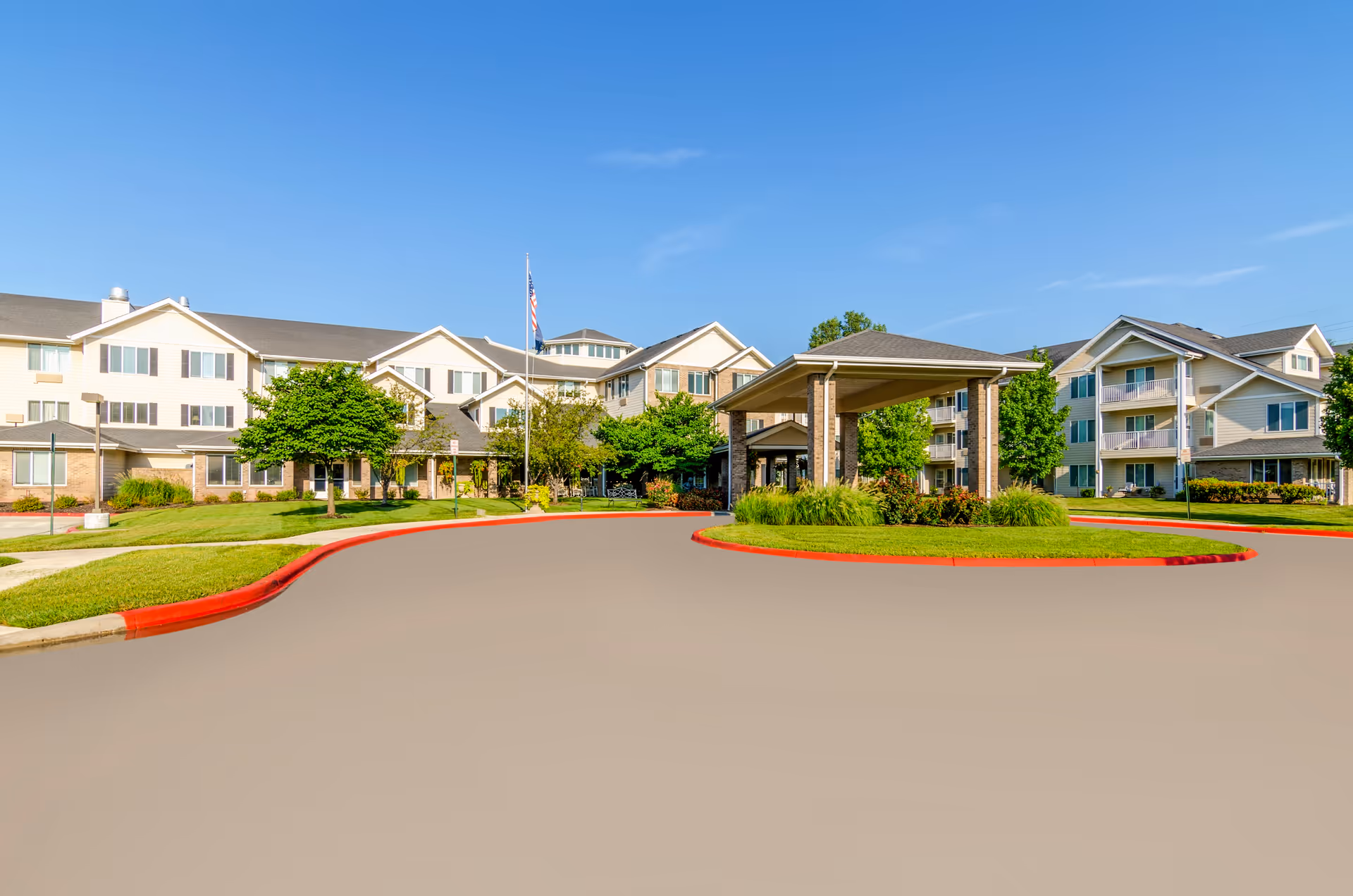 Exterior view of Holiday Greenwood Terrace, a multi-story senior living facility with beige siding and brick accents. The building is surrounded by well-maintained green lawns, trees, and shrubs. There is a covered entrance driveway with a circular driveway and an American flag flying on a flagpole in front of the building under a clear blue sky.