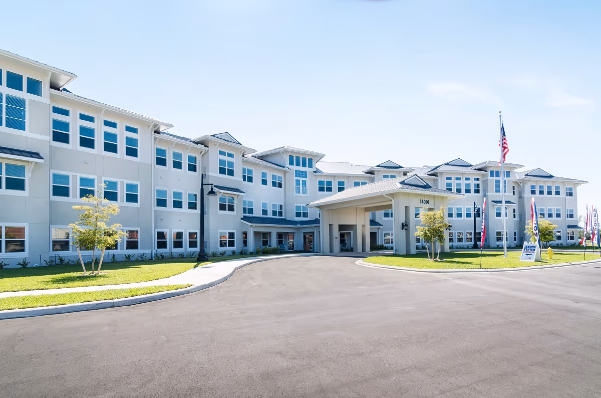 Exterior view of a large, modern three-story senior living facility building with multiple windows, a covered entrance, a driveway, and a well-maintained lawn with small trees and flagpoles displaying American flags and banners.