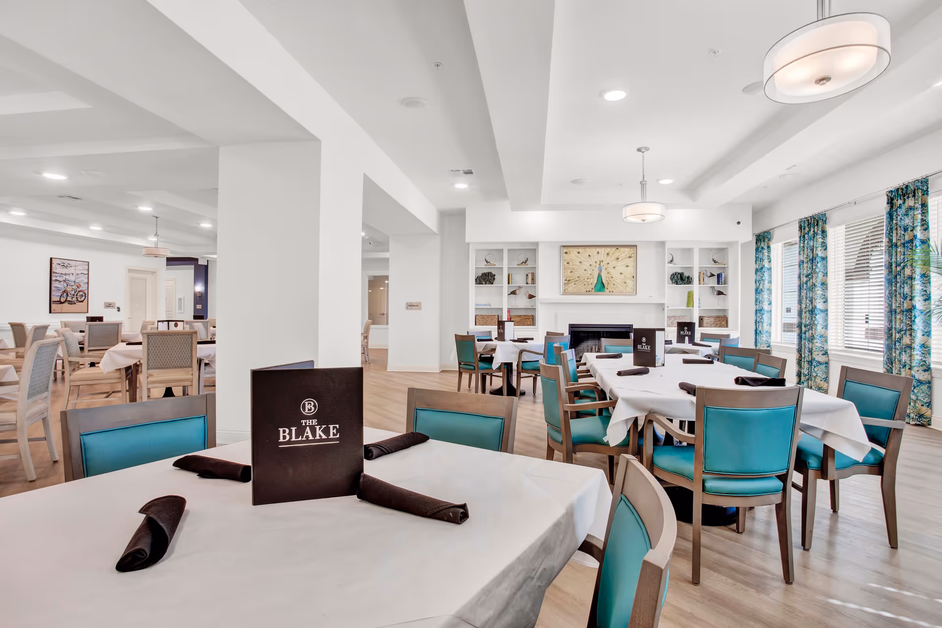 Bright dining room with white-tableclothed tables, teal chairs, and menus reading 'The Blake'.