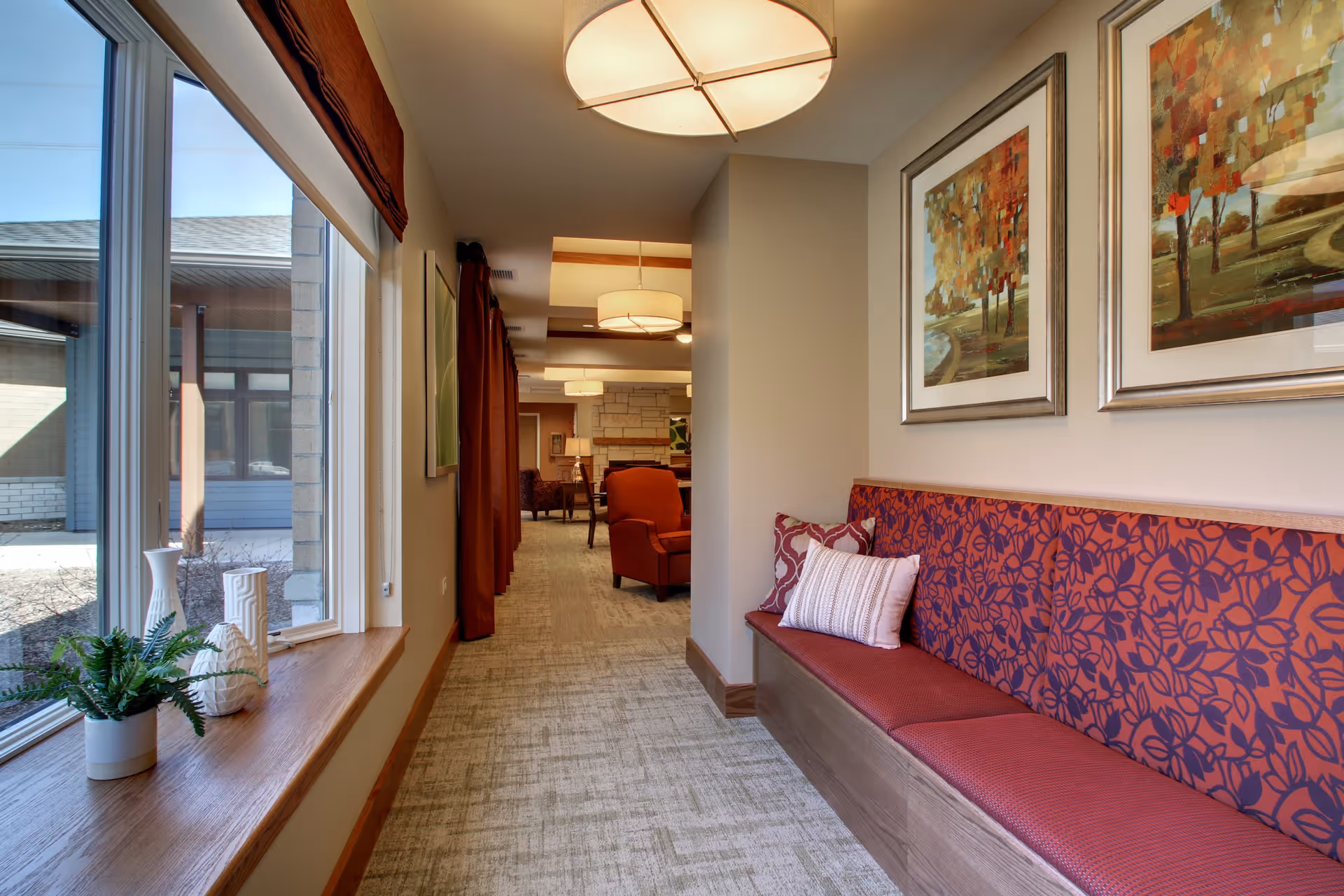 A hallway in Evergreen Senior Living with a built-in bench featuring red and purple floral cushions and pillows on the right side. Two framed paintings of autumn trees hang above the bench. On the left, large windows with wooden sills let in natural light and display outdoor views. The hallway leads to a seating area with red armchairs and a stone fireplace in the background. Ceiling lights provide warm illumination.