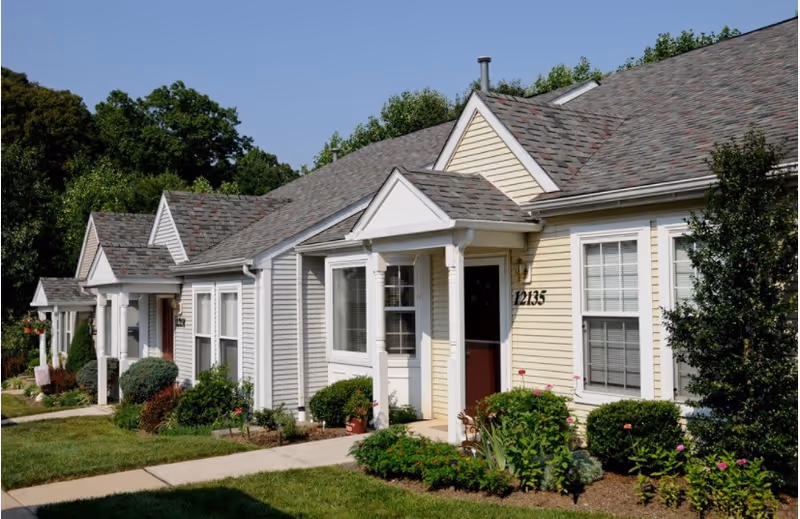 Exterior view of a row of single-story residential buildings with beige and gray siding, white trim, and small front porches surrounded by well-maintained landscaping and greenery under a clear blue sky.