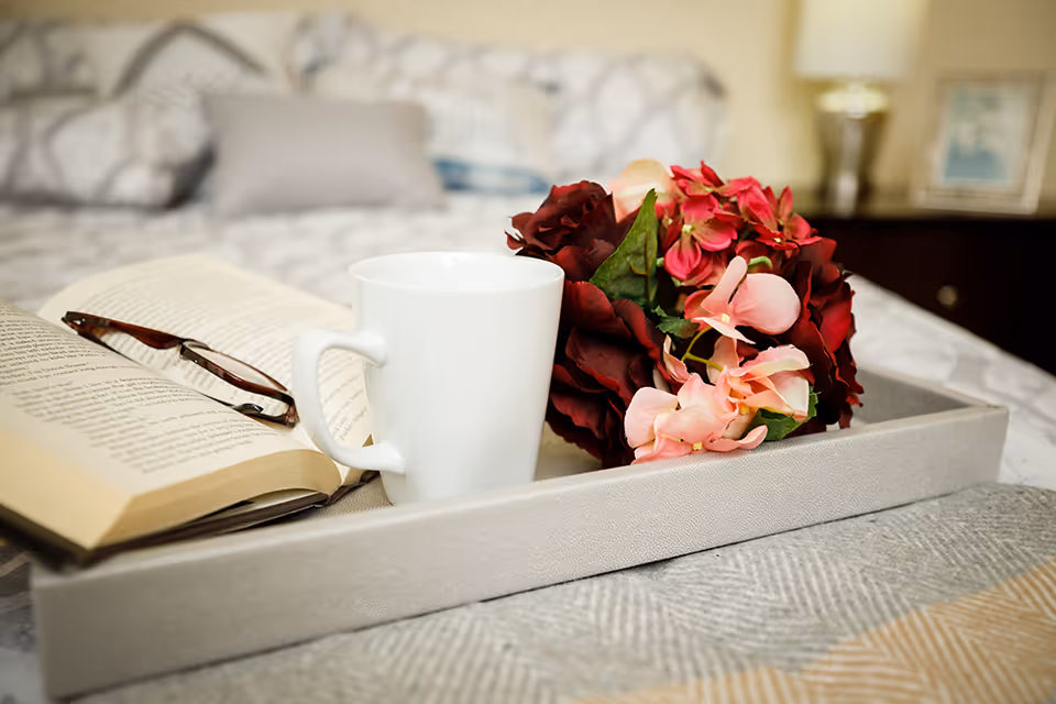 A close-up of a tray on a bed holding an open book with reading glasses resting on it, a white coffee mug, and a bouquet of red and pink flowers. The background shows a blurred bed with pillows and a nightstand with a lamp and a framed picture.