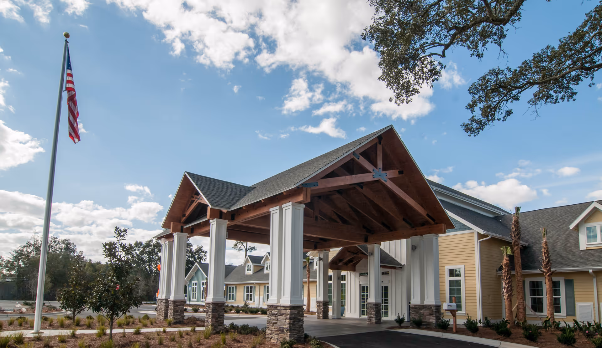 Exterior view of a senior living facility with a large covered entrance supported by white columns with stone bases. The building has yellow siding and multiple windows, with a clear blue sky and some clouds above. An American flag is flying on a flagpole to the left, and there are small landscaped plants and trees around the driveway.