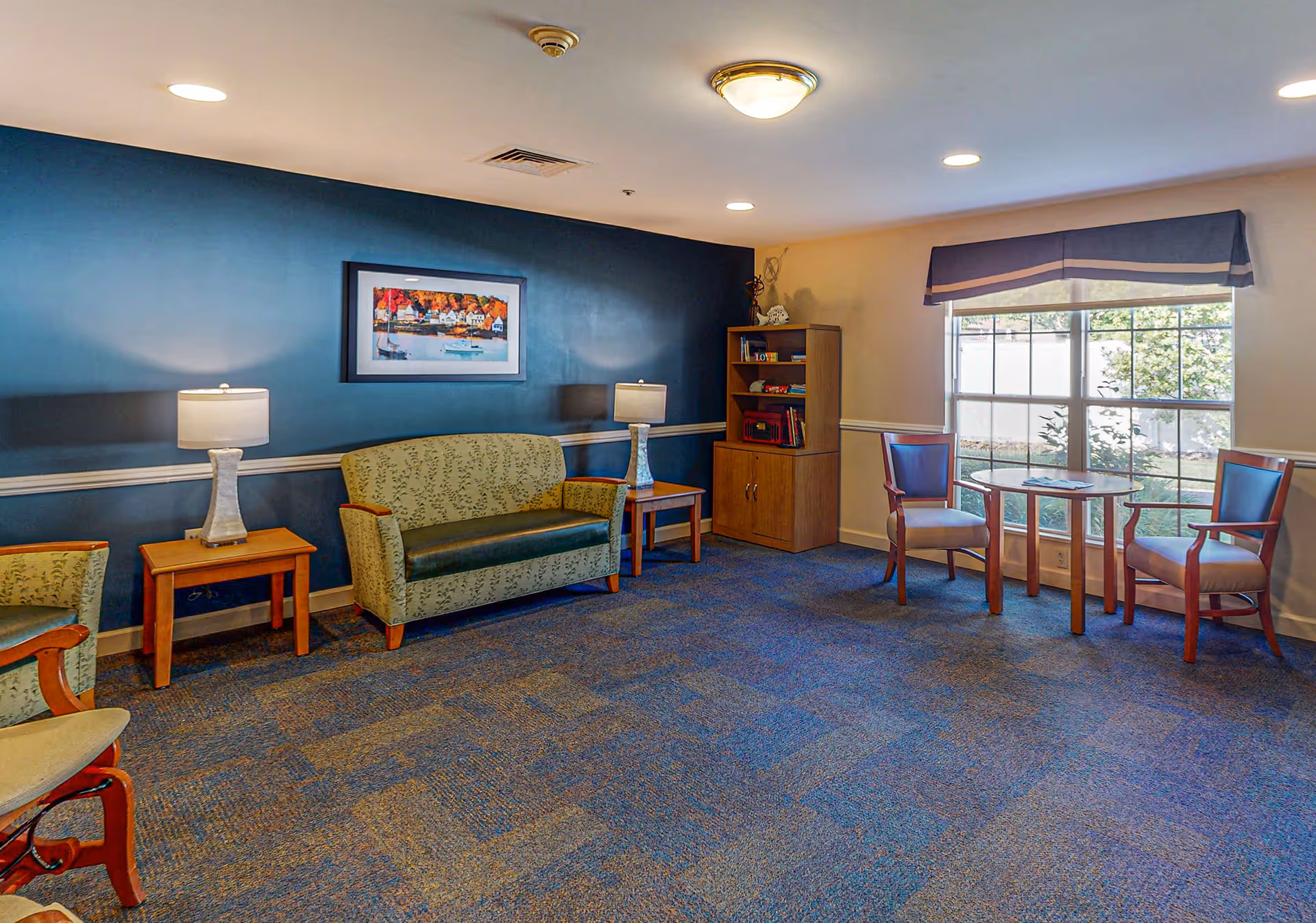 A cozy sitting area with patterned green upholstered chairs and a loveseat, two wooden side tables with lamps, a wooden bookshelf, and a round table with two chairs near a large window with a blue valance. The walls are painted blue and beige, and the carpet is a mix of blue and brown tones.