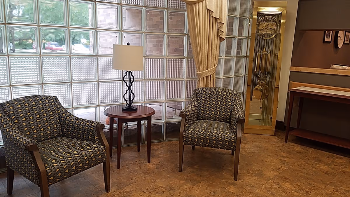A cozy sitting area with two patterned armchairs and a round wooden side table with a lamp on it. The background features a wall made of glass blocks, a tall grandfather clock, and a wooden console table against a brown wall with framed pictures.
