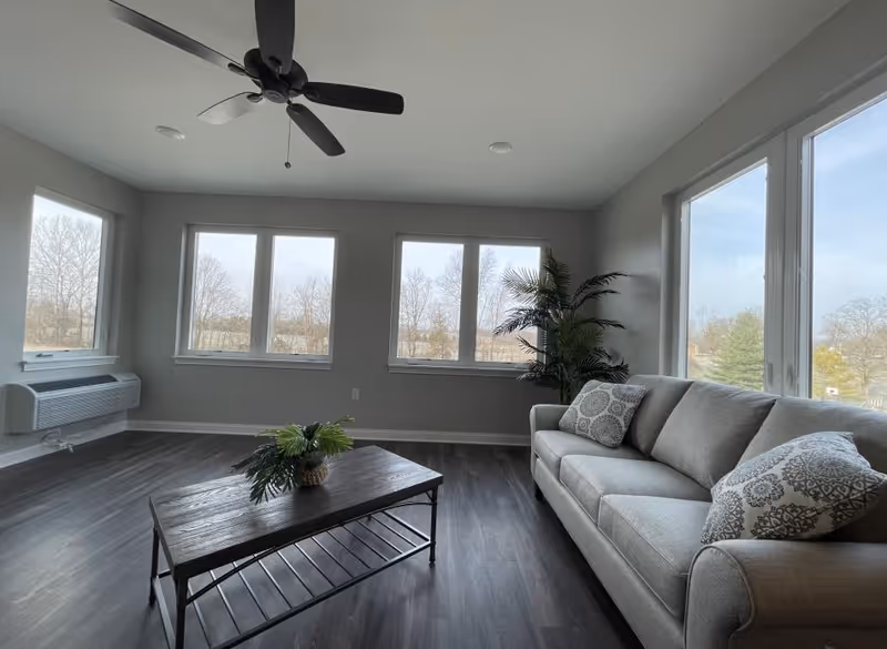 A bright living room with large windows showing a view of trees outside. The room features a gray sofa with patterned cushions, a wooden coffee table with a small plant, a ceiling fan, and a tall potted plant in the corner. The floor is dark wood, and the walls are painted light gray.